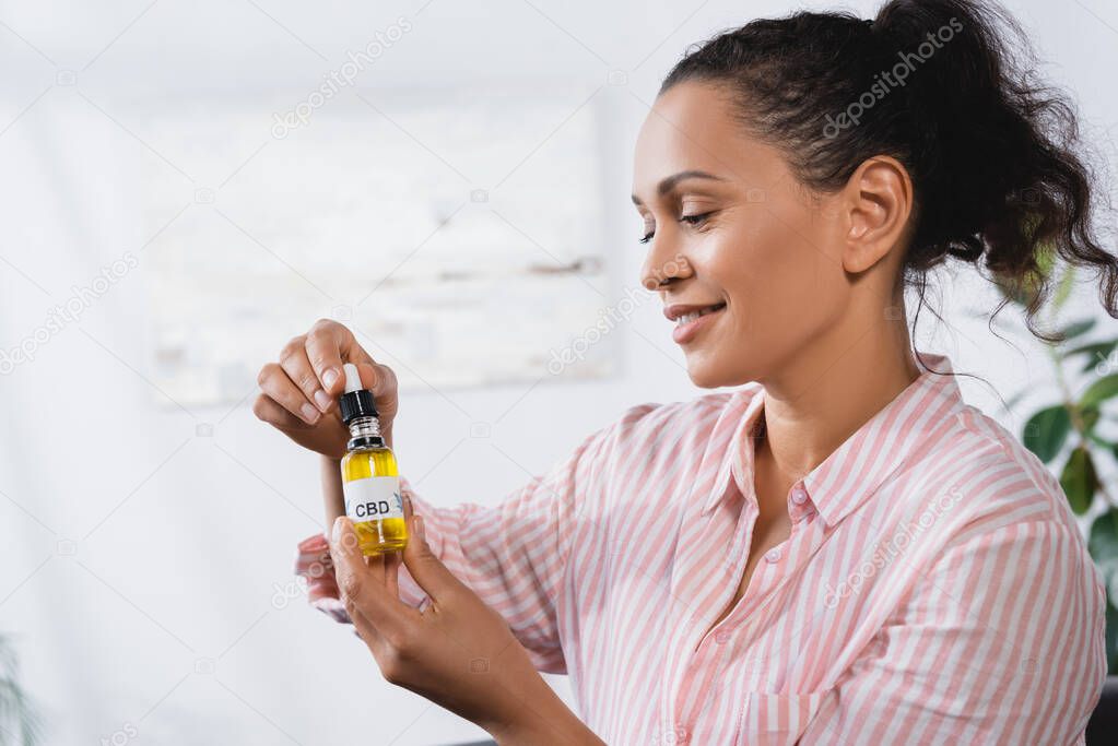 happy african american woman holding bottle with cbd lettering and pipette in living room