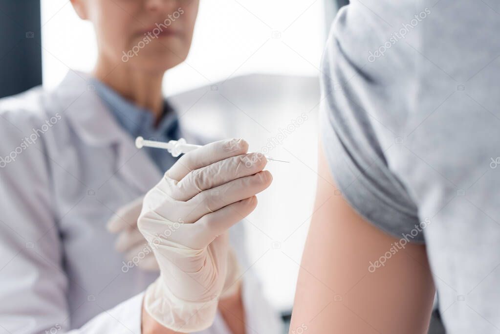 Cropped view of syringe in hand of doctor near patient on blurred foreground 