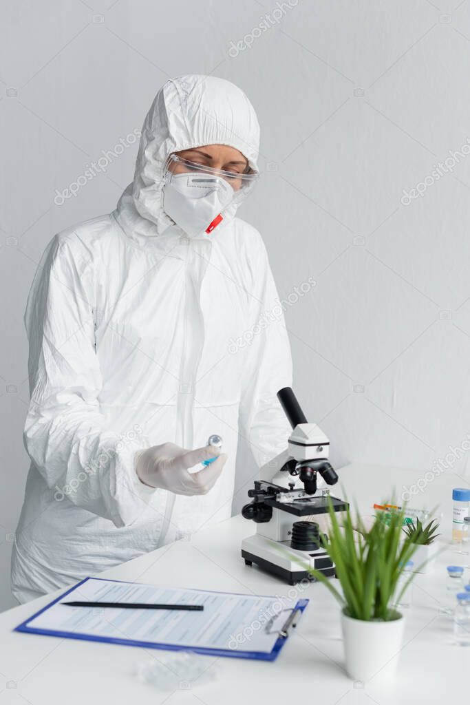 Scientist in hazmat suit looking at vaccine near clipboard and microscope in laboratory 