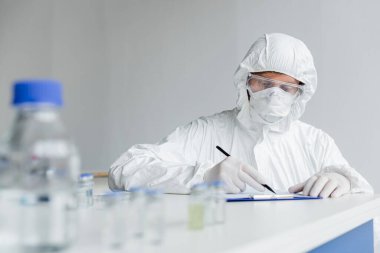 Scientist writing on clipboard near vaccines on blurred foreground on table  clipart
