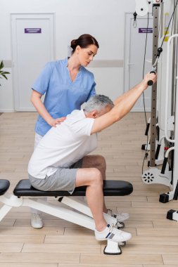 Rehabilitasyon uzmanı, olgun bir adamın hastane spor salonunda çalışmasına yardım ediyor.