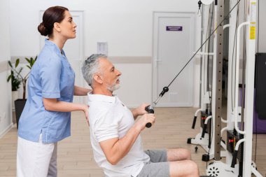 Kablo çekme makinesinde egzersiz yapan olgun bir adamı destekleyen rehabilitasyon uzmanının yan görüşü