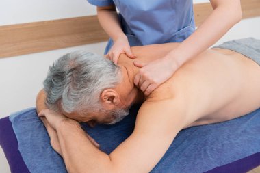 Masöz rehabilitasyon merkezinde orta yaşlı bir adama sırt masajı yapıyor.