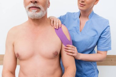 Rehabilitasyon uzmanının, yetişkin bir adamın omzuna Kinesio bandı yapıştırması.