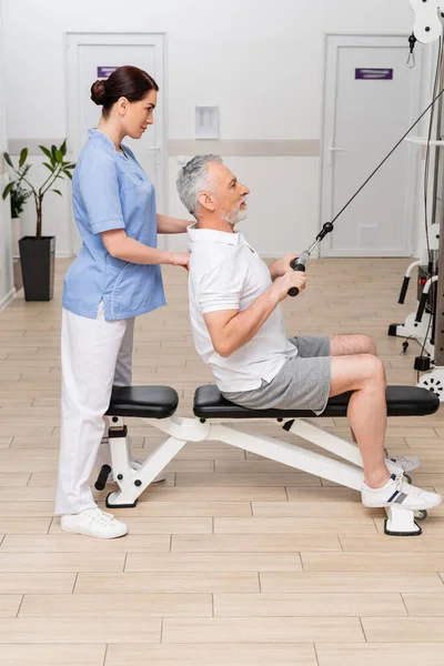 side view of physical instructor supporting mature man during rehabilitation in hospital gym