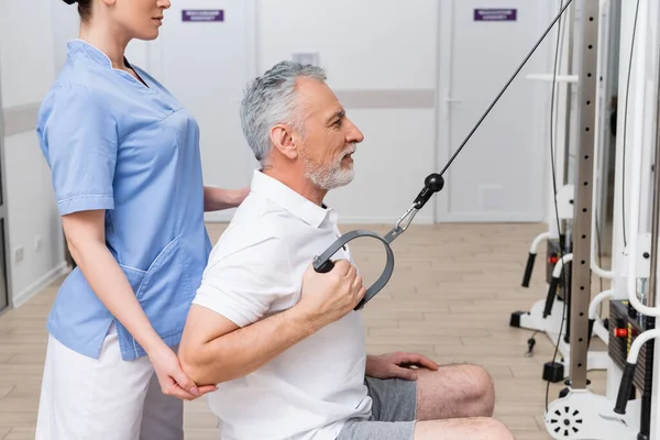 physical instructor assisting mature man working out on pull cable exercising machine