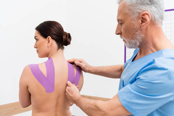 mature rehabilitologist applying tape on back of woman during kinesiotherapy