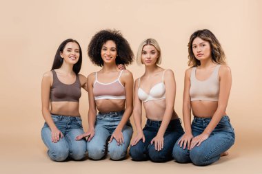 Seksi ırklar arası kadınlar kameraya bakarken bej renkli sütyen ve kot pantolon giyerler.