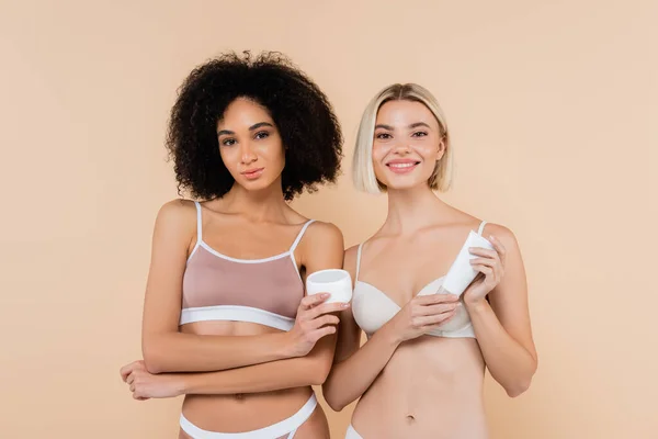 Pretty Multiethnic Friends Holding Cosmetic Cream While Smiling Camera Isolated — Stock Photo, Image