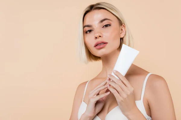 pretty woman holding tube of cosmetic cream while looking at camera isolated on beige