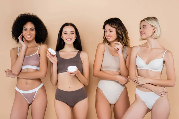 happy woman holding body cream near multicultural friends on beige