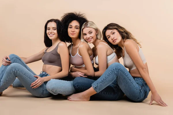 Joyful Interracial Women Smiling Camera While Sitting Bras Jeans Beige — Stock Photo, Image