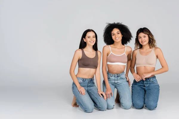 Feliz Interracial Las Mujeres Sujetadores Jeans Mirando Cámara Gris — Foto de Stock