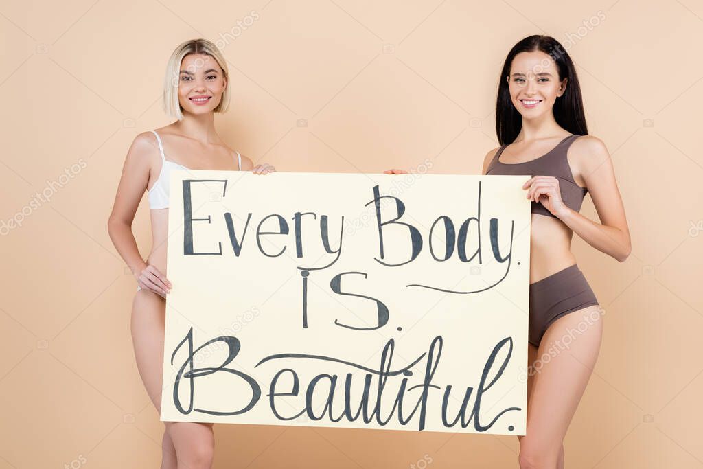 smiling women in underwear holding placard with every body is beautiful lettering on beige
