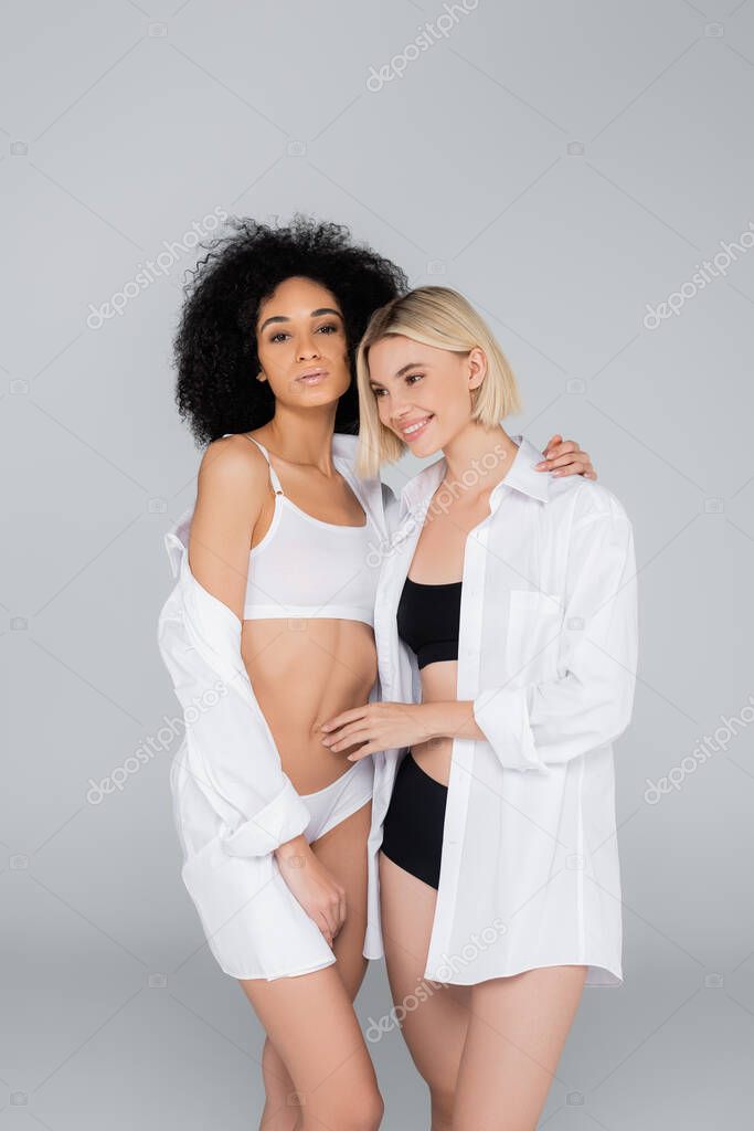 pretty multiethnic woman in white shirts and underwear embracing isolated on grey