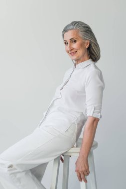 Gri saçlı kadın kameraya gülümsüyor. Gri sandalyede izole edilmiş. 