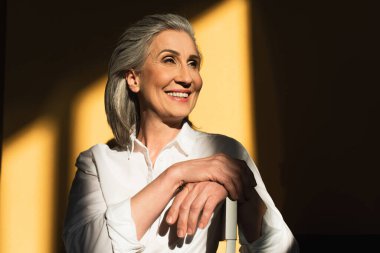 Gülümseyen olgun kadın, sarı arka plandaki sandalyeye bakıyor. 