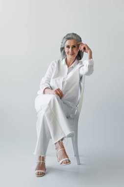 Gülümseyen olgun bir kadın gri arka planda bacak bacak üstüne atmış bir şekilde oturuyor. 
