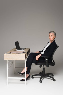 Mature businesswoman sitting near gadgets and papers on table on grey background  clipart