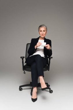 Resmi olarak iş kadını, gri arka plandaki ofis sandalyesinde elinde bir fincan kahve tutuyor. 
