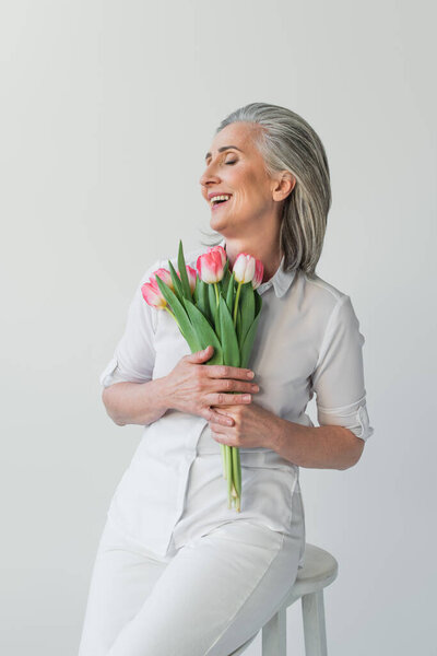 Mature woman with closed eyes smiling while holding tulips isolated on grey 