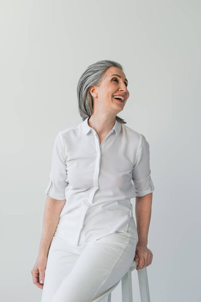 Grey haired woman in white shirt looking away on chair isolated on grey 