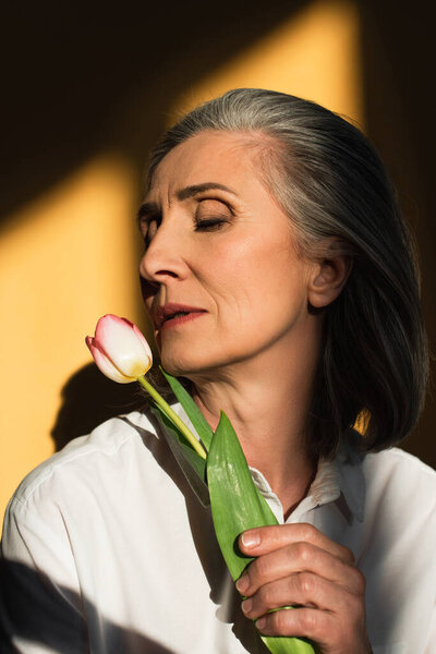 Mature woman in white shirt holding tulip on yellow background with shadow 