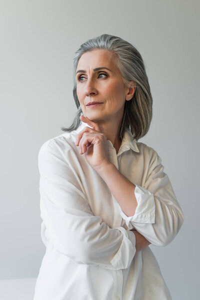 Mature woman with hand near chin posing isolated on grey 