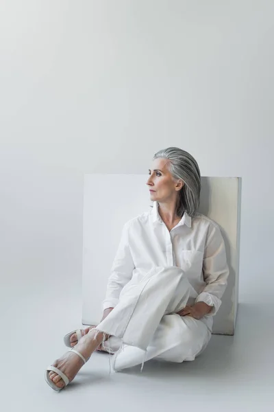 Grey haired woman sitting near white cube on grey background