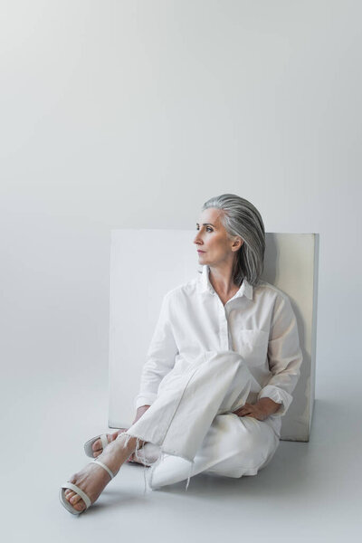Grey haired woman sitting near white cube on grey background 
