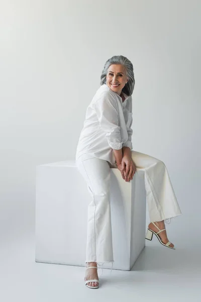 Smiling Mature Woman Sitting White Cube Grey Background — Stock Photo, Image