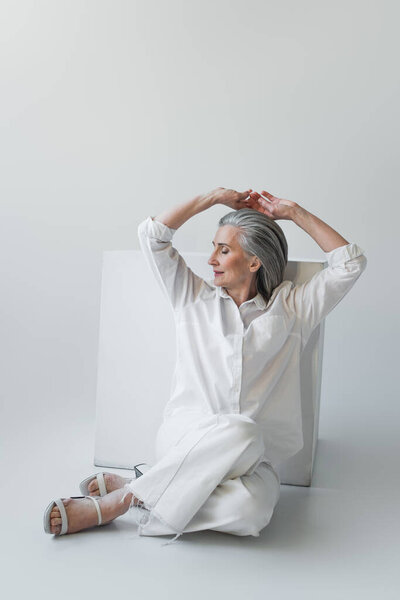 Grey haired woman posing near white cube on grey background 