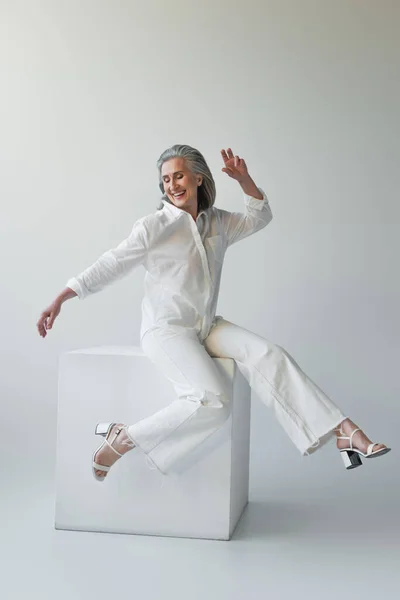 Happy mature woman sitting on cube on grey background