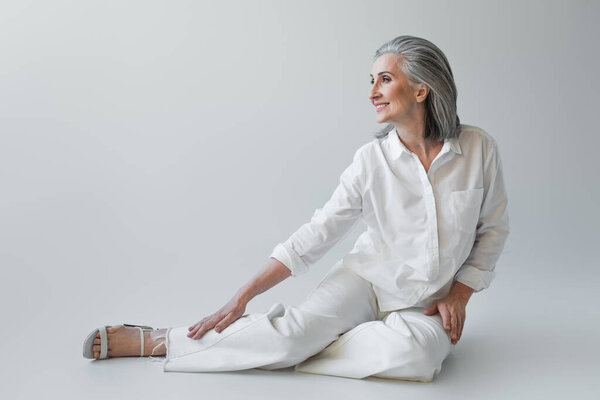 Cheerful mature woman in white clothes sitting on grey background 