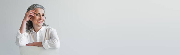 Grey haired woman smiling at camera while sitting on chair isolated on grey, banner 