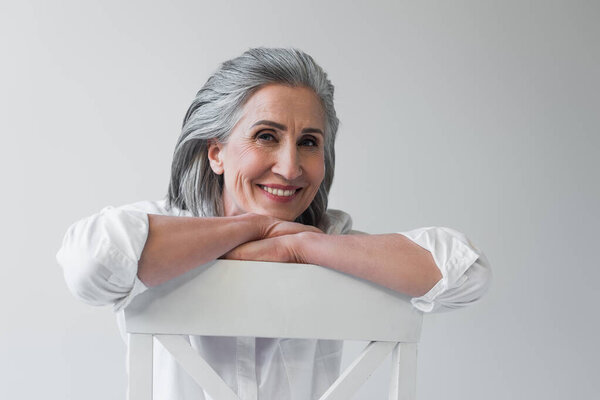 Happy grey haired woman looking at camera on chair isolated on grey 