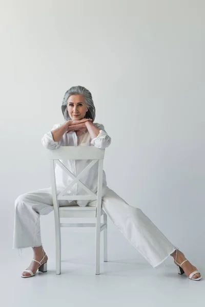 一个成熟的女人坐在灰色背景的椅子上看着相机 — 图库照片