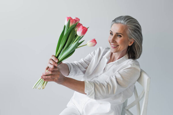 Mature woman smiling while looking at tulips on chair isolated on grey 
