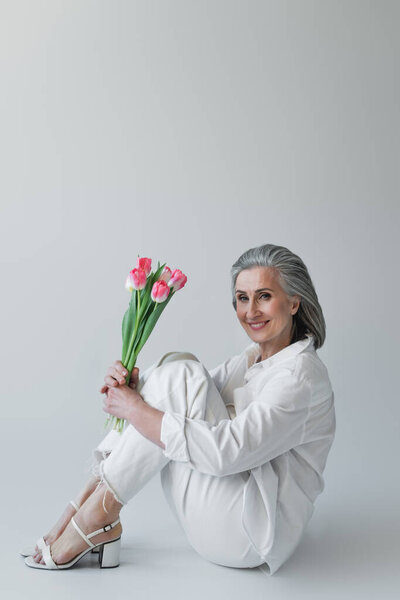 Middle aged woman holding flowers and smiling at camera while sitting on grey background 