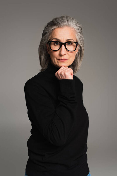 Зрелая женщина в черном свитере и очках, смотрящая на камеру, изолированную на сером 