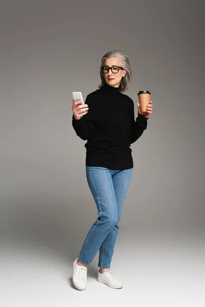 在灰色背景下使用智能手机和拿着外卖饮料的成熟女人 — 图库照片