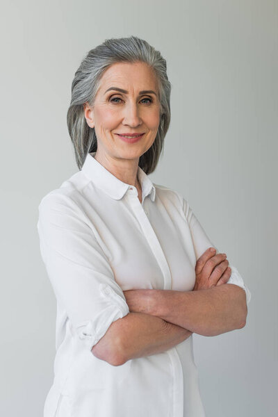 Улыбающаяся зрелая женщина со скрещенными руками, изолированная на сером 