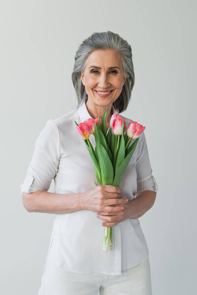 Positive mature woman in white shirt holding tulips isolated on grey 