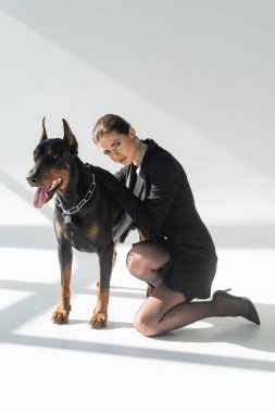 Şehvetli, esmer kadın gölgeli gri arka planda doberman köpeğine yaslanıyor.