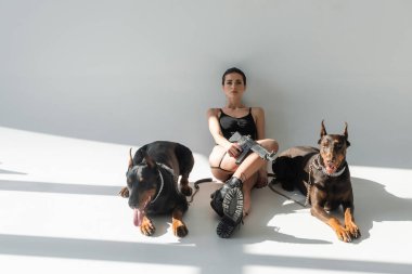 Siyah elbiseli ve deri çizmeli silahlı bir kadın gri arka planda gölgeli Dobermanlar 'ın yanında oturuyor.