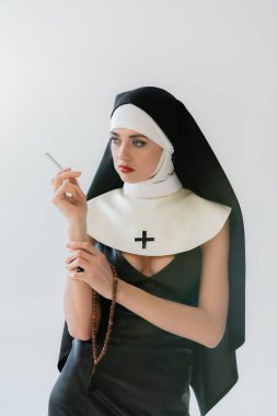 seductive nun looking away while smoking isolated on grey clipart