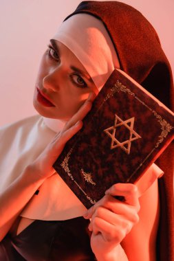 young, sexy nun looking at camera while holding jewish bible isolated on pink clipart