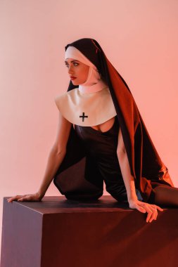 young nun in sexy dress looking away while sitting on black cube isolated on pink clipart