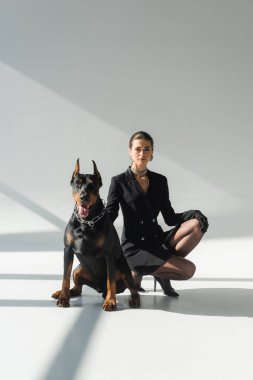 Gri arka planda gölgeli doberman köpeğinin yanında siyah ceketli zarif bir kadın.