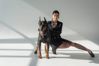 Gri arka planda gölgeli doberman köpeğinin yanında poz veren siyah ceketli şık bir kadın.